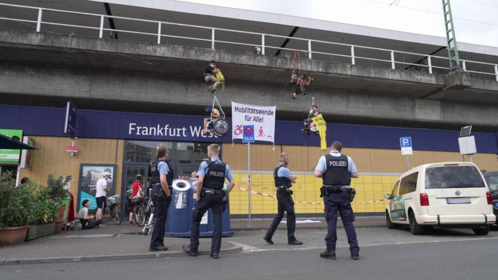 Vor dem Eingang zum Frankfurt West Bahnhof hängen vier Menschen auf Kletterseile, zwei davon im Rollstuhl, mit einem großen Banner "Mobilitätswende für Alle!". Vier Polizist*innen stehen etwas ratlos davor.