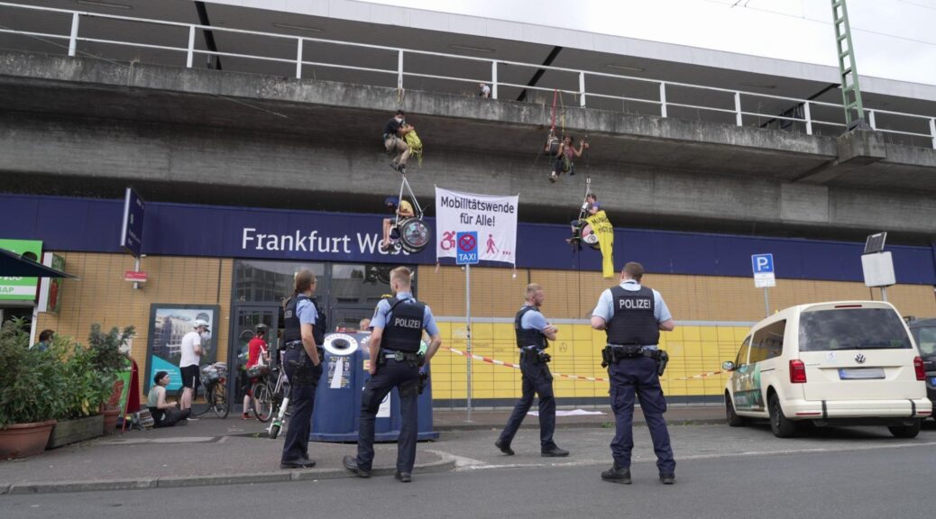Four people hang from climbing ropes in front of the entrance to Frankfurt West train station. Two of them are in wheelchairs, and between them is a big banner saying "Mobility transition for everyone!" Four police officers are standing on the ground below them.
