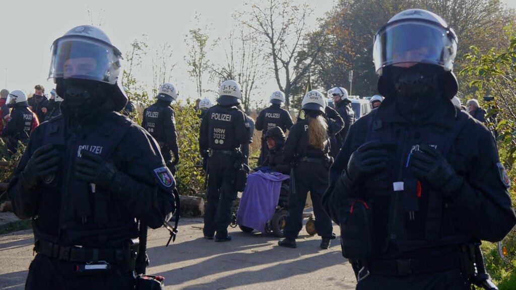 A person in an electric wheelchair blocking a road, encircled by many police officers in riot gear.