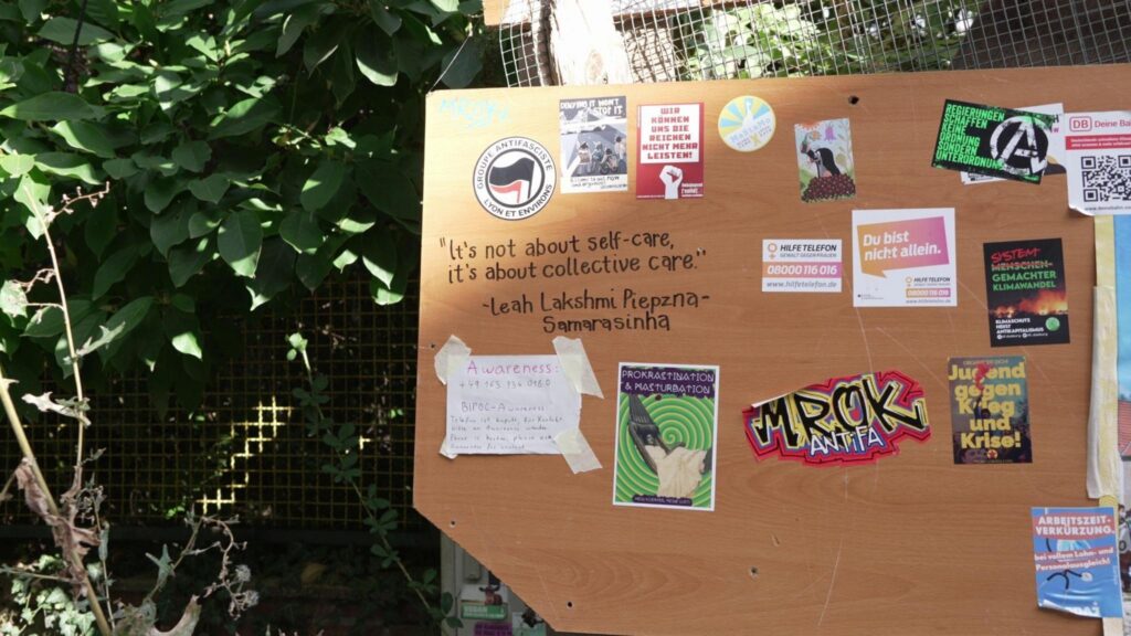 A big board attached to a fence, with political stickers. Written in marker pen is a quote "It's not about self-care, it's about collective care" from Leah Lakshmi Piepzna-Samarasinha.