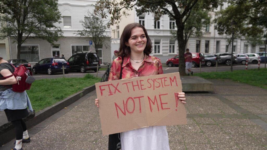 A person with long brown hair and a white skirt holds a sign: "Fix the system not me!"
