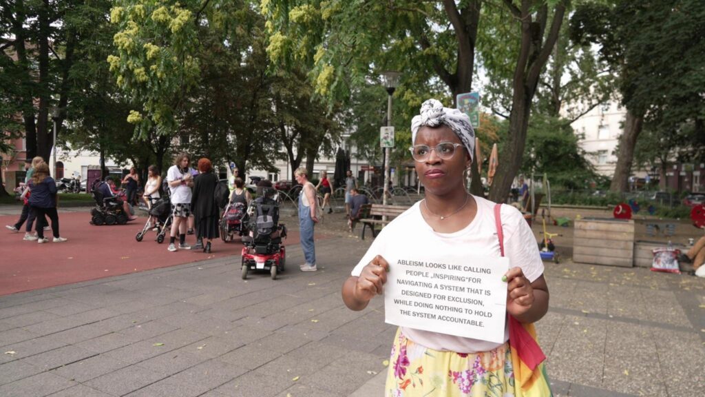 Tabitha, a colourfully dressed Black artist with a rainbow bag holds a sign: "Ableism looks like calling people 'inspiring' for navigating a system that is designed for exclusion, while doing nothing to hold the system accountable".