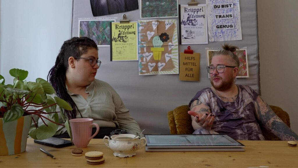 SchwarzRund and simo tier sit at a table and talk. Behind them are posters and zines on the wall: "Mobility aids for everyone!", "You are trans enough", "Cripple Newspaper" (all translated from German)