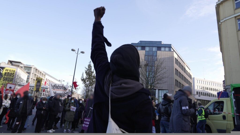 A person in a demonstration holds their left fist high in the air. In front of them a loudspeaker van and lots of people with banners and signs. "Justice for Mouhamed" is readable.