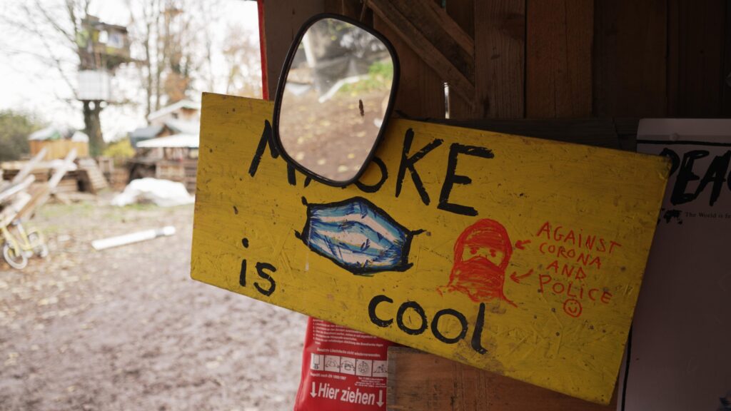 yellow painted sign that reads" Mask is cool, against Corona and Police :)", in the background a treehouse